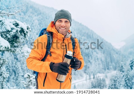 Laughing Man dressed bright orange softshell jacket with a hot drink thermos flask looking at camera while he trekking winter mountains route. Active people in the nature concept image. Royalty-Free Stock Photo #1914728041