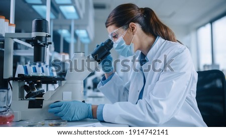 Female Scientist in Face Mask and Glasses Looking a Petri Dish with Genetically Modified Sample Chemicals Under a Microscope. Microbiologist Working in Modern Laboratory with Technological Equipment. Royalty-Free Stock Photo #1914742141