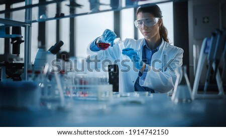 Beautiful Female Scientist Wearing Protective Goggles Mixing Chemicals in a Test Tube in a Lab. Young Professional Microbiologist Working in Modern Laboratory with Technological Equipment. Royalty-Free Stock Photo #1914742150