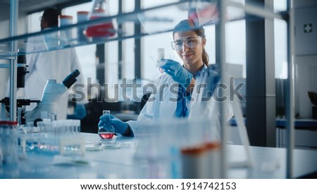 Beautiful Female Scientist Wearing Protective Goggles Mixing Chemicals in a Test Tube in a Lab. Young Professional Microbiologist Working in Modern Laboratory with Technological Equipment. Royalty-Free Stock Photo #1914742153