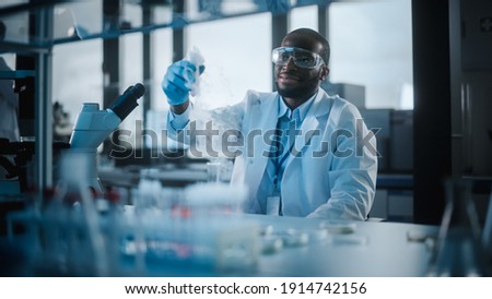 African American Male Scientist Wearing Protective Goggles Mixing Chemicals in a Test Tube in a Lab. Handsome Black Microbiologist Working in Modern Laboratory with Technological Equipment. Royalty-Free Stock Photo #1914742156