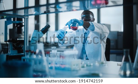 African American Male Scientist Wearing Protective Goggles Mixing Chemicals in a Test Tube in a Lab. Handsome Black Microbiologist Working in Modern Laboratory with Technological Equipment. Royalty-Free Stock Photo #1914742159