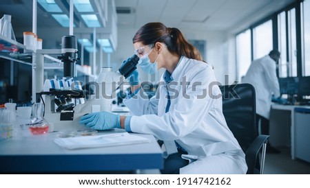 Female Scientist in Face Mask and Glasses Looking a Petri Dish with Genetically Modified Sample Chemicals Under a Microscope. Microbiologist Working in Modern Laboratory with Technological Equipment. Royalty-Free Stock Photo #1914742162