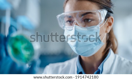 Beautiful Female Scientist Wearing Face Mask and Glasses Looking a Petri Dish with Genetically Modified Sample Chemicals. Microbiologist Working in Modern Laboratory with Technological Equipment. Royalty-Free Stock Photo #1914742180