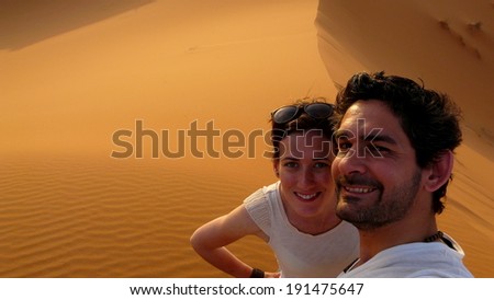 A young couple taking a self picture while climbing to the top of the Great Sand Dune in the red dune sea of Erg Chebbi, Morocco. 