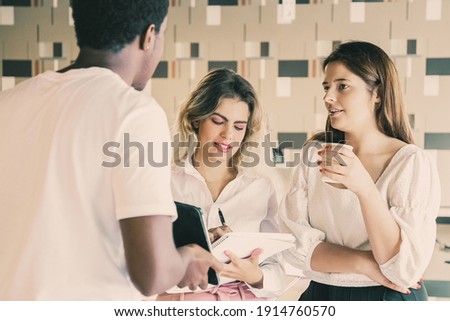 African American guy showing tablet screen to two women. Young long-haired Caucasian colleagues standing, making notes and listening designer. Communication, digital technology and teamwork concept