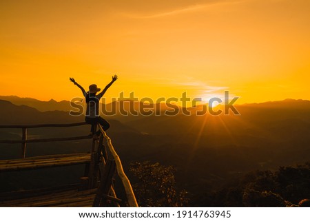 Picture from the back of a woman sitting and raise both arm on a wooden porch extending into a high mountain cliff. The sun is setting on the mountain and there is a beautiful warm orange light.