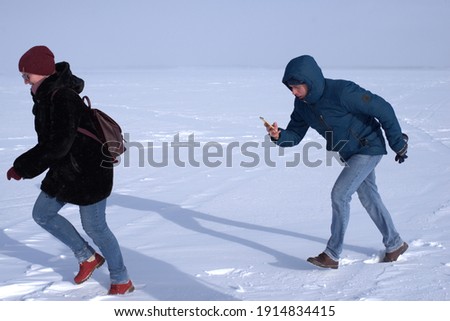 A girl in a burgundy hat, brown fur coat, blue jeans and red shoes runs in the snow. A young man in a blue jacket and jeans runs after the girl and takes her on a smartphone. Sunny day on a snowy lake