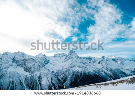 Photo of mountain peaks in sunny weather in winter close-up. Snowy mountains, white clouds, blue sky. 