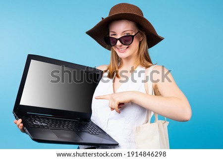 A beautiful blonde girl with sunglasses on her head and hat points to a laptop monitor. Blue background . High quality photo