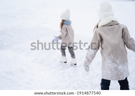 Beautiful little girls, sisters learn to skate on ice skating rink in park. Fall and have fun. Stylish looks, warm woolen coats, white hats, scarves, snoods. Winter family activities, games outdoors.