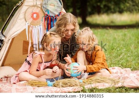 Mom is spending time with her little daughters, teaching them geography in a playful way. Girls have native americans make up on their faces. Wigwam decoration with dream catchers in the background. 