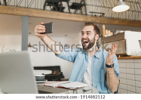 Excited man sit alone at table in coffee shop cafe restaurant indoors working studying on laptop pc computer doing selfie shot on cell phone spreading hands. Freelance mobile office business concept