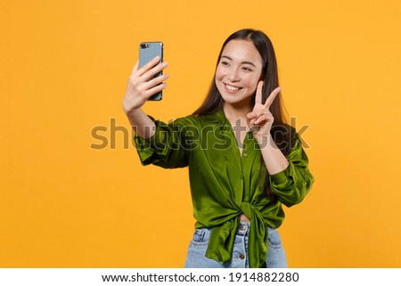 Smiling cheerful young brunette asian woman wearing basic green shirt standing doing selfie shot on mobile phone showing victory sign isolated on bright yellow colour background, studio portrait