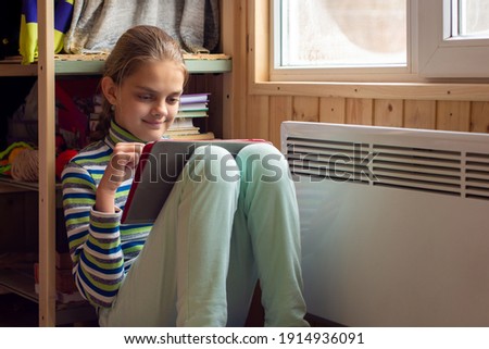 A girl plays in a tablet sitting next to a radiator by a window in a country house