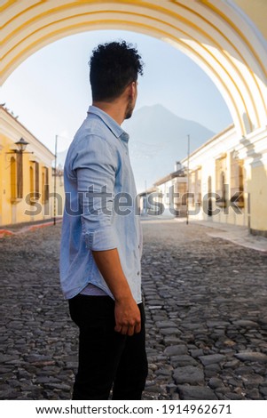 Hispanic young man standing under the Arch of Santa Catalina in Antigua Guatemala - tourist on important avenue of colonial Guatemala city with the Water volcano background