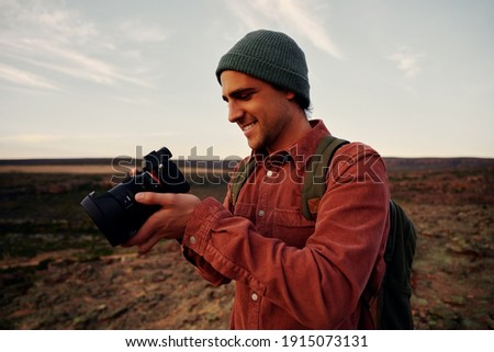 Male travel photographer checking photos on camera after shot standing on mountain wearing cap and backpack
