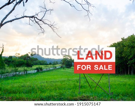 Land for sale sign on empty land, green meadow near the tree and surrounded with white wooden fence on beautiful sky background. Red and white letters on sign board. Real estate concept.