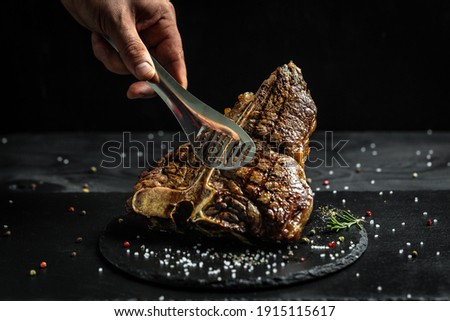 chef hand holding steak meat tongs. large piece of fresh beef meat prepared on a grill. Medium rare Grilled T-Bone Steak, Barbecue aged wagyu porterhouse. Royalty-Free Stock Photo #1915115617