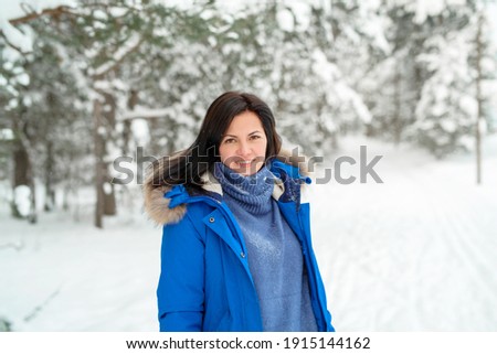 A young brunette woman in a blue jacket stands in the snow and watching into the camera in a winter forest.