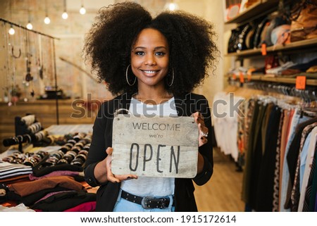 Portrait of the owner of the clothing store holding the sign with the words "Welcome we're open" in her hand to be attached to the window - Millennial woman welcomes customers at the entrance