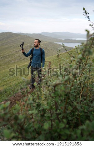Tourist man with camera and backpack takes a photo on his phone standing on the hill Green bushes Grey sky