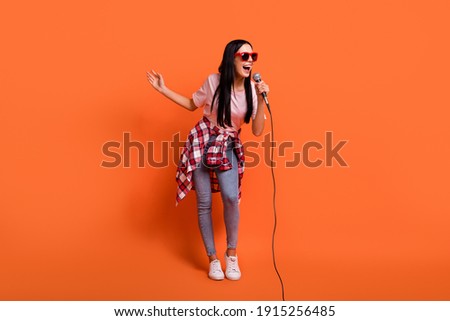 Full size photo of optimistic brunette lady stand sing in mic wear spectacles t-shirt jeans isolated on orange background Royalty-Free Stock Photo #1915256485