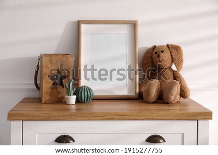 Empty photo frame near cute toy bunny and decor on dresser, space for text. Baby room interior element