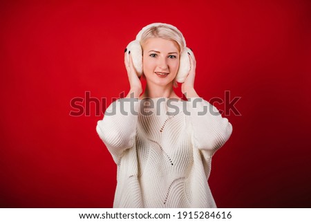 A young woman in a white knitted sweater and fur headphones stands on a red background. A girl with a cute smile in winter clothes. Studio photo