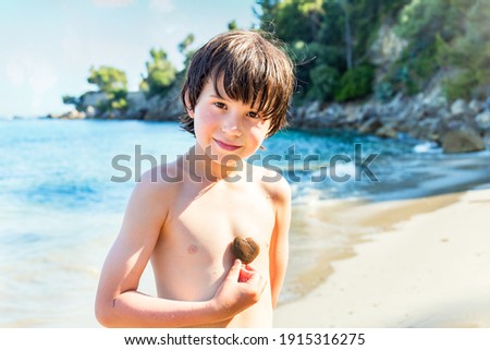Young boy spending time at the seaside keeping heart-shaped pebble stone at his chest. Smiling child in summer at the beach during holidays.