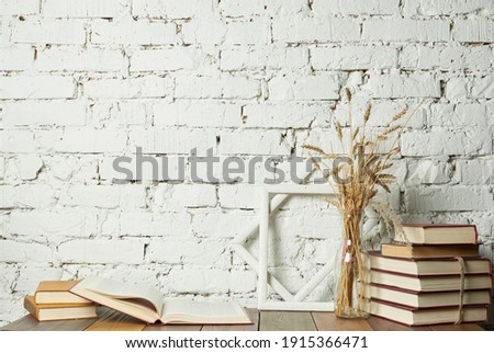 Wooden shelf with stack of books, wooden frames and dry spikelets in glass vase against white brick wall background