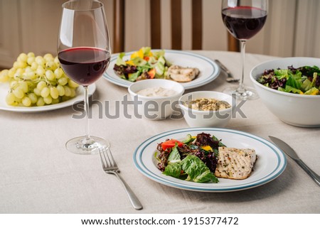 White fish steak with vegetable salad, sauce, hummus, grapes and glasses of wine. Served for two in the kitchen