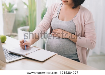 Closeup of a pregnant woman writing notes and using a laptop while working on maternity leave at her dining room table at home Royalty-Free Stock Photo #1915384297