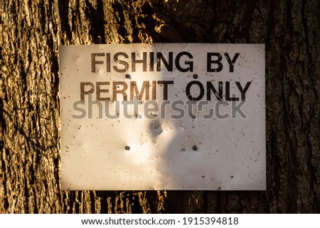 Close up of a fishing by permit only sign attached to a large tree in the winter sun Royalty-Free Stock Photo #1915394818