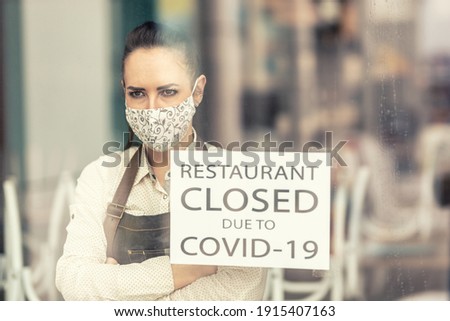Upset restaurant owner stands with arms crossed on her chest next to a sign saying restaurant closed due to covid-19.