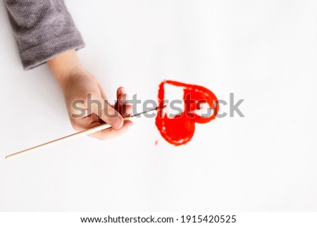 A child holds a brush in his hands and draws a red heart on white paper, close-up top view. Postcard for the holiday, Valentine's Day, Mother's Day, International Women's Day.