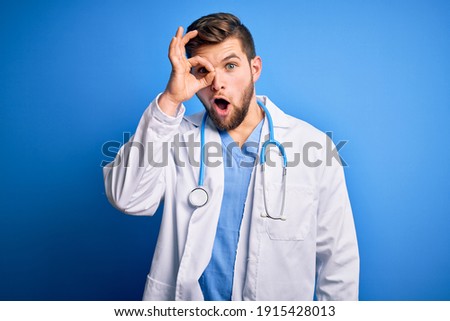 Young blond doctor man with beard and blue eyes wearing white coat and stethoscope doing ok gesture shocked with surprised face, eye looking through fingers. Unbelieving expression.