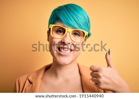 Young beautiful woman with blue fashion hair wearing casual glasses over yellow background happy with big smile doing ok sign, thumb up with fingers, excellent sign