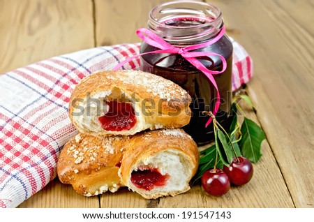 Puff bun with jam, a jar of cherry marmalade, cherries with leaves, a napkin on a wooden boards background