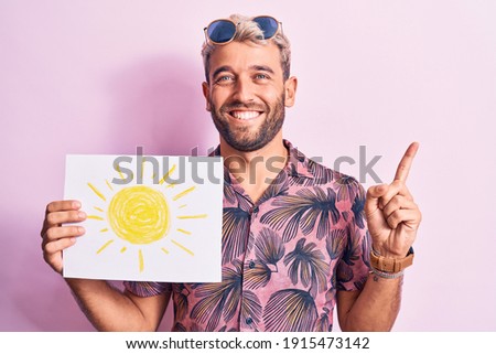 Handsome blond tourist man on vacation wearing sunglasses holding paper with sun draw smiling happy pointing with hand and finger to the side