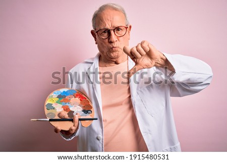 Senior grey haired artist man painting using painter palette over pink background with angry face, negative sign showing dislike with thumbs down, rejection concept
