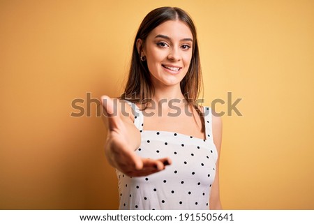 Young beautiful brunette woman wearing casual dress standing over yellow background smiling friendly offering handshake as greeting and welcoming. Successful business.
