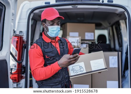 Young african delivery man with package and smartphone wearing surgical face mask for coronavirus outbreak Royalty-Free Stock Photo #1915528678