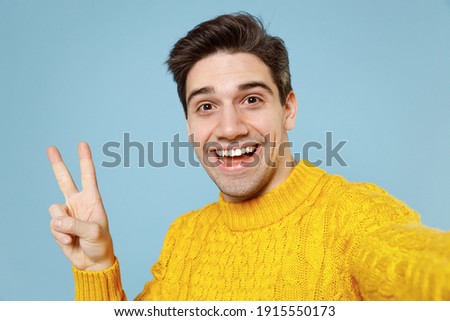 Close up young caucasian happy smiling student man 20s in knitted yellow fashionable sweater doing selfie shot on mobile phone show v-sign victory gesture isolated on blue background studio portrait.