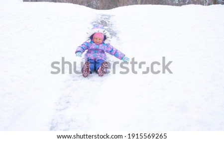 A little pretty girl in a purple overalls slides down an ice slide, winter children's games, a child in the park in winter.
