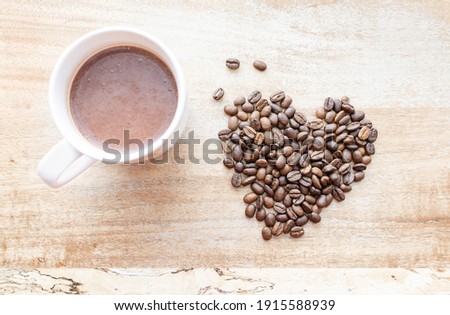 Top view of  Roasted brown coffee beans formed in a heart shape near a cup of hot beverage on wood background, depth of field. The aromatic idea for coffee lovers, a symbol of love on Valentine’s Day.