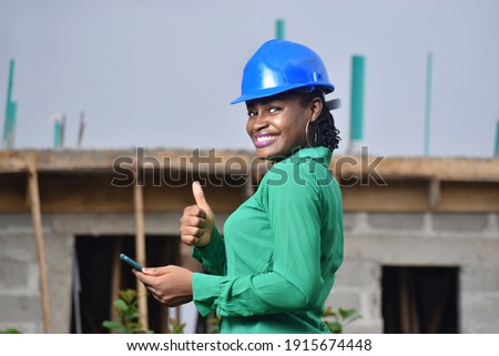 A Nigerian female construction and architectural engineer with blue safety helmet holding a phone and giving thumbs up  Royalty-Free Stock Photo #1915674448
