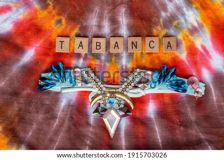 Tabanca, spelled out in wooden letters, with a bedazzled blue and pink diamond costume headpiece from Trinidad and Tobago carnival. Multicoloured tie dye fabric background.