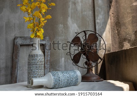 Yellow bouquet of flowers in White ceramic vase bottle shape and Vintage brown metal fan on white textured table cloth with old cement wall at the balcony house. Home decor, Selective focus.
