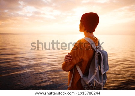 A charming calm young woman stands by the sea expressing delight. Hipster woman traveler with backpack in yellow sweater and red hat enjoying sunset on the coast.